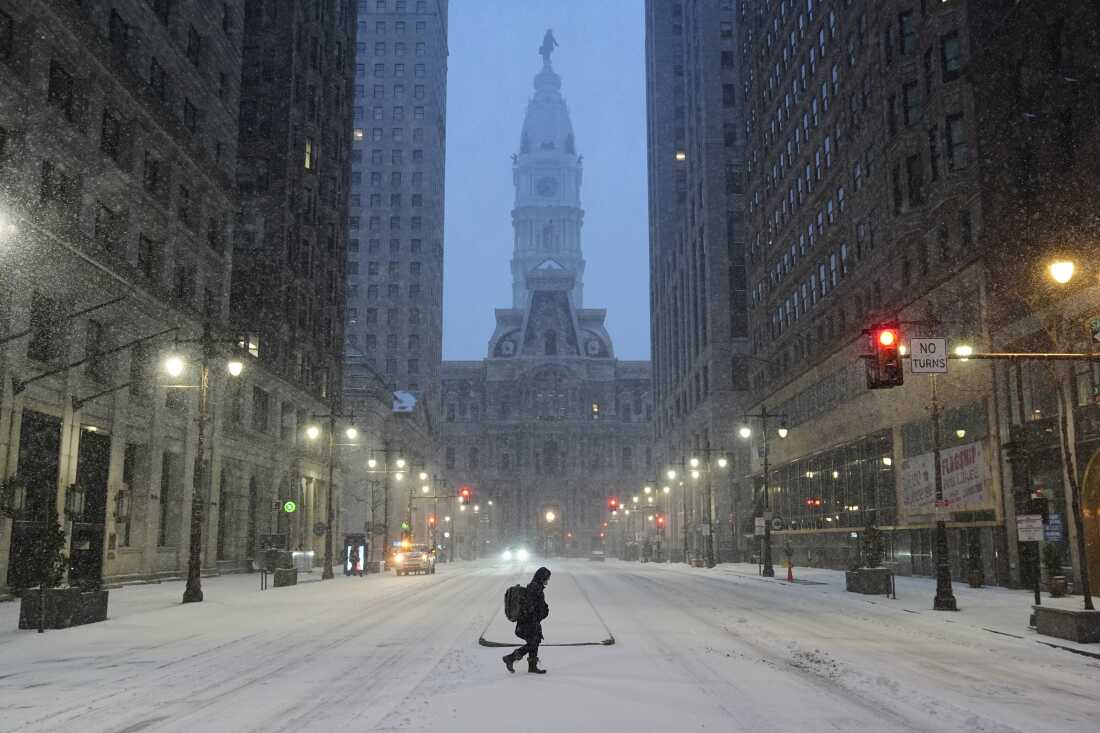 Le 25 janvier, une personne portant un manteau et des bottes traverse une rue enneigée lors d'une tempête hivernale à Philadelphie. De grands immeubles s'élèvent des deux côtés de la photo.