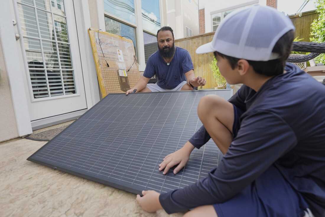 Accroupis près du sol à l'extérieur, Bhavin Misra et son fils Rumi tiennent les côtés opposés d'un panneau solaire carré noir avec un motif en grille tout en assemblant un kit solaire enfichable chez eux à Houston.