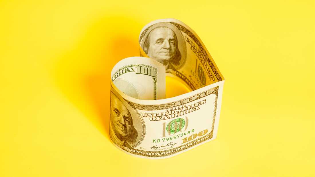 Deux billets de 100 dollars posés sur leurs bords longs sont disposés pour former ensemble une forme de cœur. Ils sont sur une surface jaune.