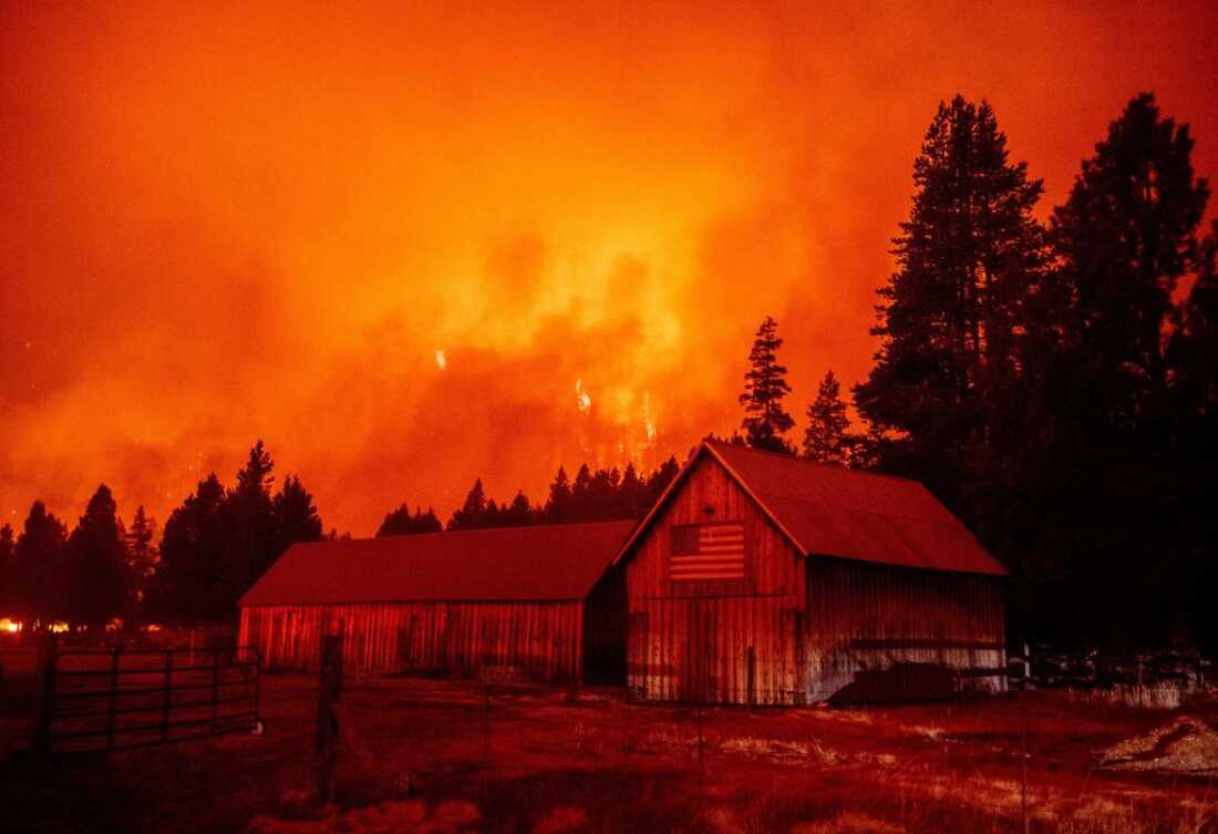 Des flammes, de la fumée et un ciel orange forment la toile de fond d’une structure avec un drapeau américain sur le côté et de grands arbres derrière.