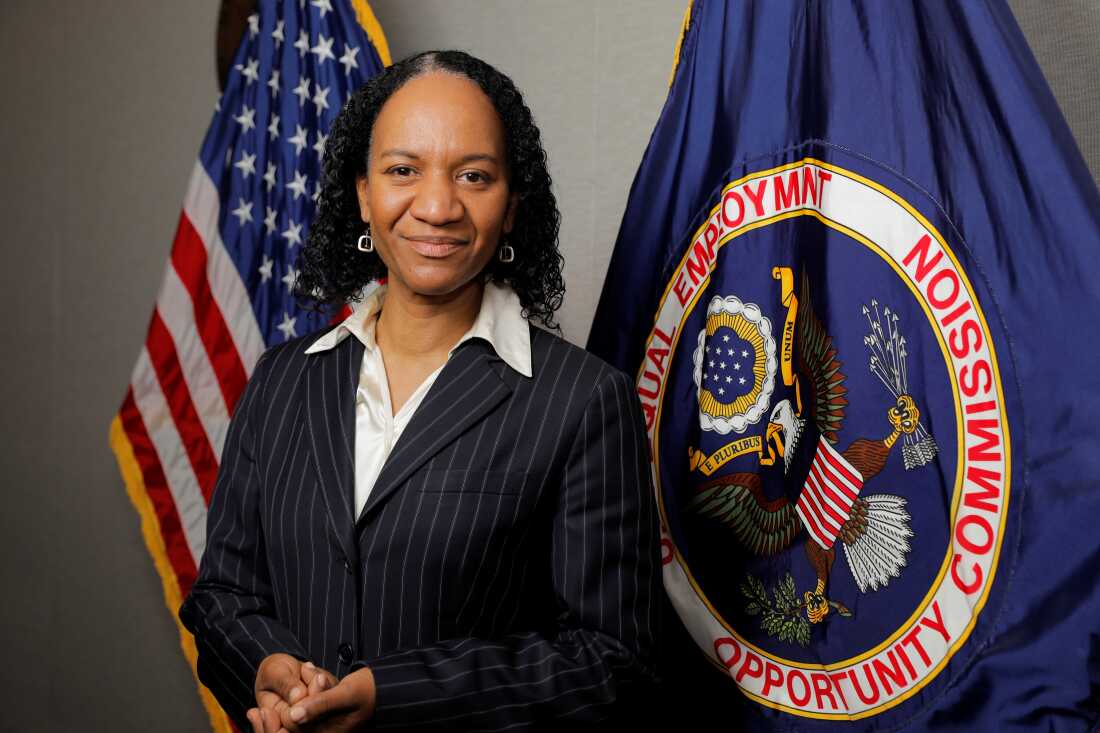 Charlotte Burrows, alors présidente de l'EEOC, en 2021. C'est une femme noire debout devant un drapeau américain et le drapeau de l'EEOC, qui est bleu et sur lequel figure un aigle.