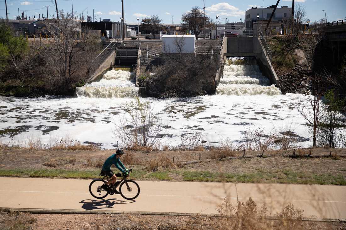Un cycliste roule à vélo sur un chemin qui passe par des émissaires d'eaux usées traitées d'un blanc mousseux qui se jettent dans la rivière South Platte. L'installation de traitement de Metro Water Recovery à Denver est en arrière-plan.