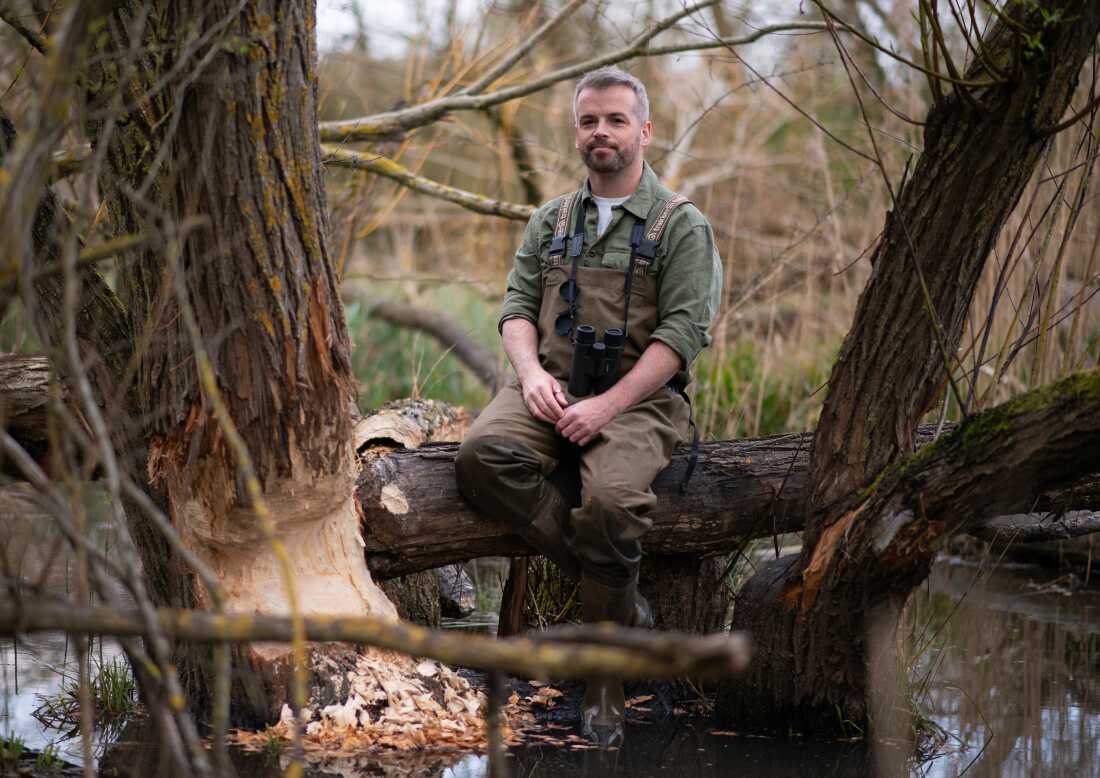 Sur cette photo, Sean McCormack est assis sur une bûche dans une zone marécageuse près d'un arbre dont le tronc a été rongé par des castors. Il porte des jumelles autour du cou et des cuissardes style salopette.