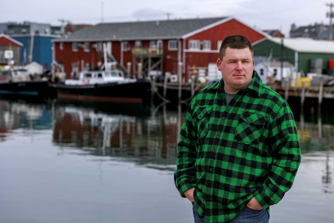 Le pêcheur Devyn Campbell se tient près de l'eau, à l'extérieur du Portland Fish Exchange. Des bateaux et une marina sont derrière lui.