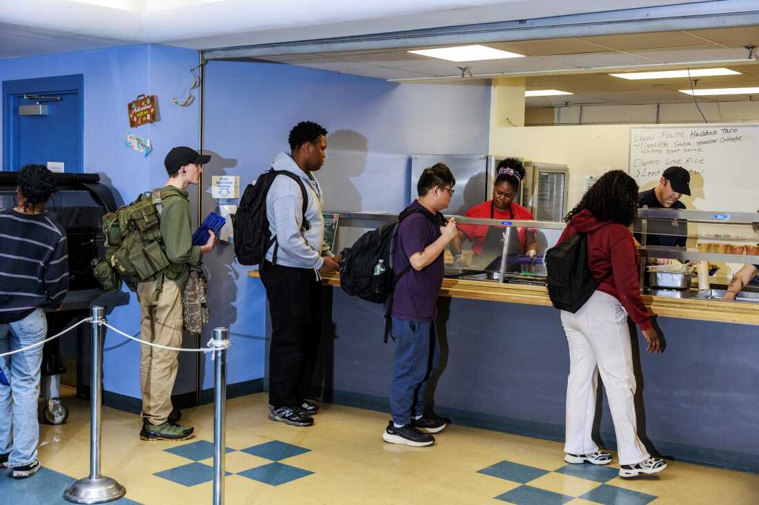 Cinq élèves du lycée Westbrook font la queue à la cafétéria pour le déjeuner. Les employés de la cafétéria se tiennent de l’autre côté d’une cloison transparente et préparent la nourriture dans des récipients métalliques.
