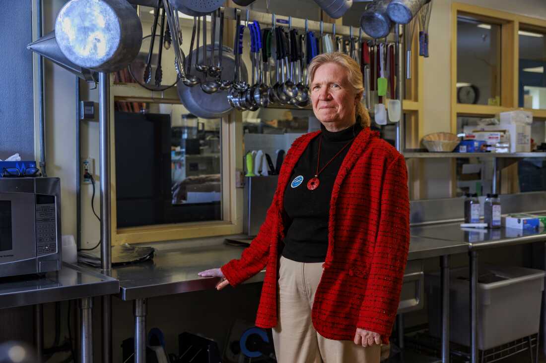 Mary Emerson se tient dans la cuisine de l'école. Derrière elle se trouve une surface de préparation de nourriture en métal brillant, et de grandes cuillères et louches sont suspendues à une tige métallique au-dessus.