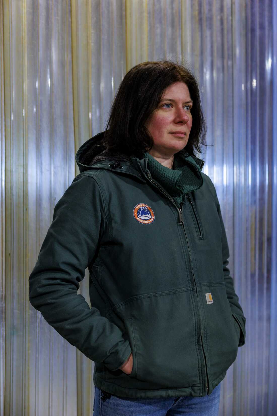 Mary Hudson se tient devant un mur en plastique. Elle porte une veste verte et un jean bleu.