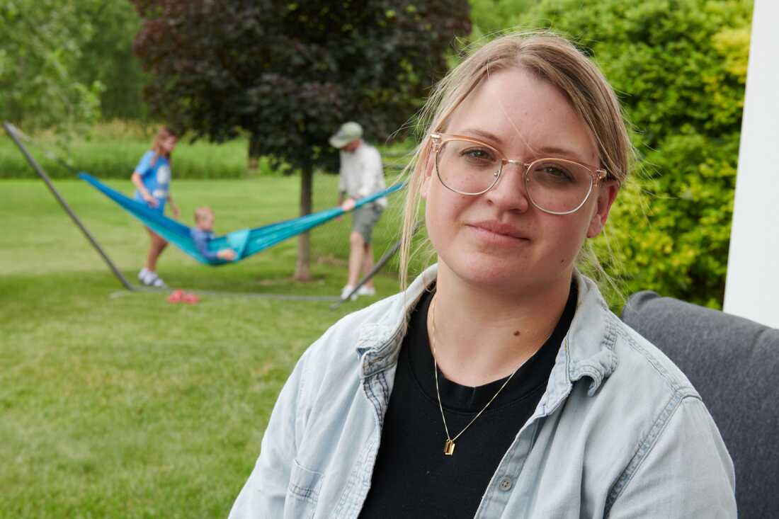 Amber Petersen est assise dans le jardin de la maison de ses parents à Iowa City, Iowa. Elle a les cheveux blonds tirés en arrière et porte des lunettes. Son mari et ses enfants jouent dans un hamac derrière elle.