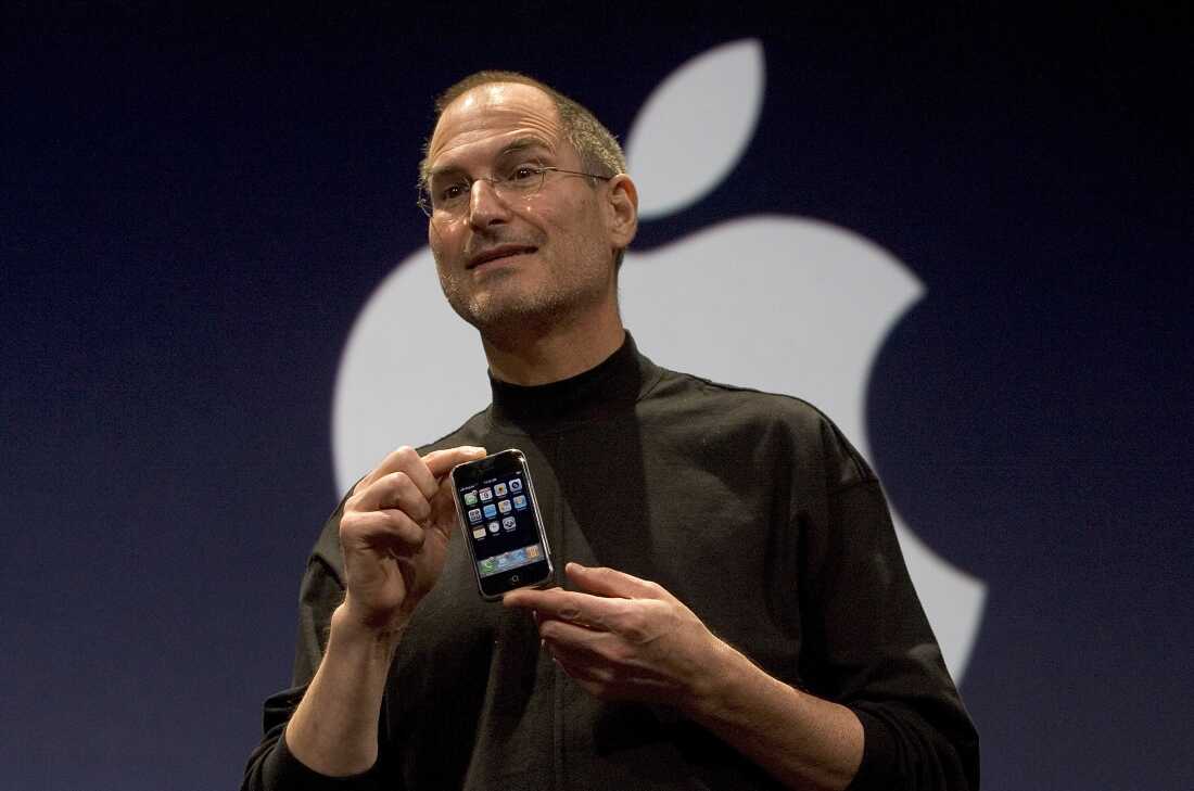 Sur cette photo de 2007, Steve Jobs, alors PDG d'Apple, tient un iPhone lors du dévoilement du smartphone. Jobs porte une chemise à col roulé noire et se tient devant un fond noir affichant le logo Apple en blanc.