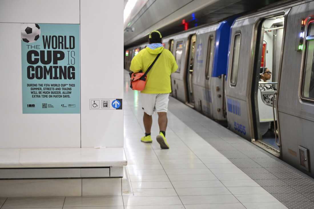 Une affiche bleu clair qui dit "La Coupe du monde approche" est affiché sur un mur blanc de la gare PATH du World Trade Center à New York le 30 mai. Un train métallique argenté se trouve sur le côté droit du cadre et un homme, dos à la caméra, marche le long du quai.