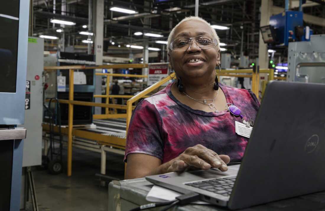 Ruth Ransom, 68 ans, est une femme aux cheveux blancs courts. Elle porte des lunettes de sécurité et se tient derrière un ordinateur portable dans l'usine.