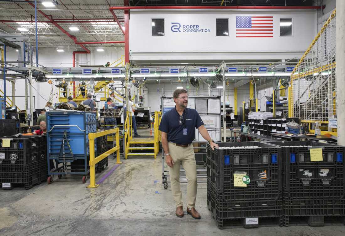 Tony Gabbert est debout dans l'usine, portant des lunettes de sécurité, un polo bleu marine et un pantalon kaki. Les ouvriers vaquent à leur travail derrière lui.