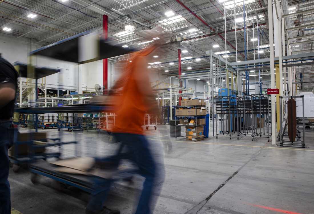 Un ouvrier, vêtu d'un gilet orange et photographié comme un flou mobile, tire un chariot derrière lui alors qu'il traverse l'usine.