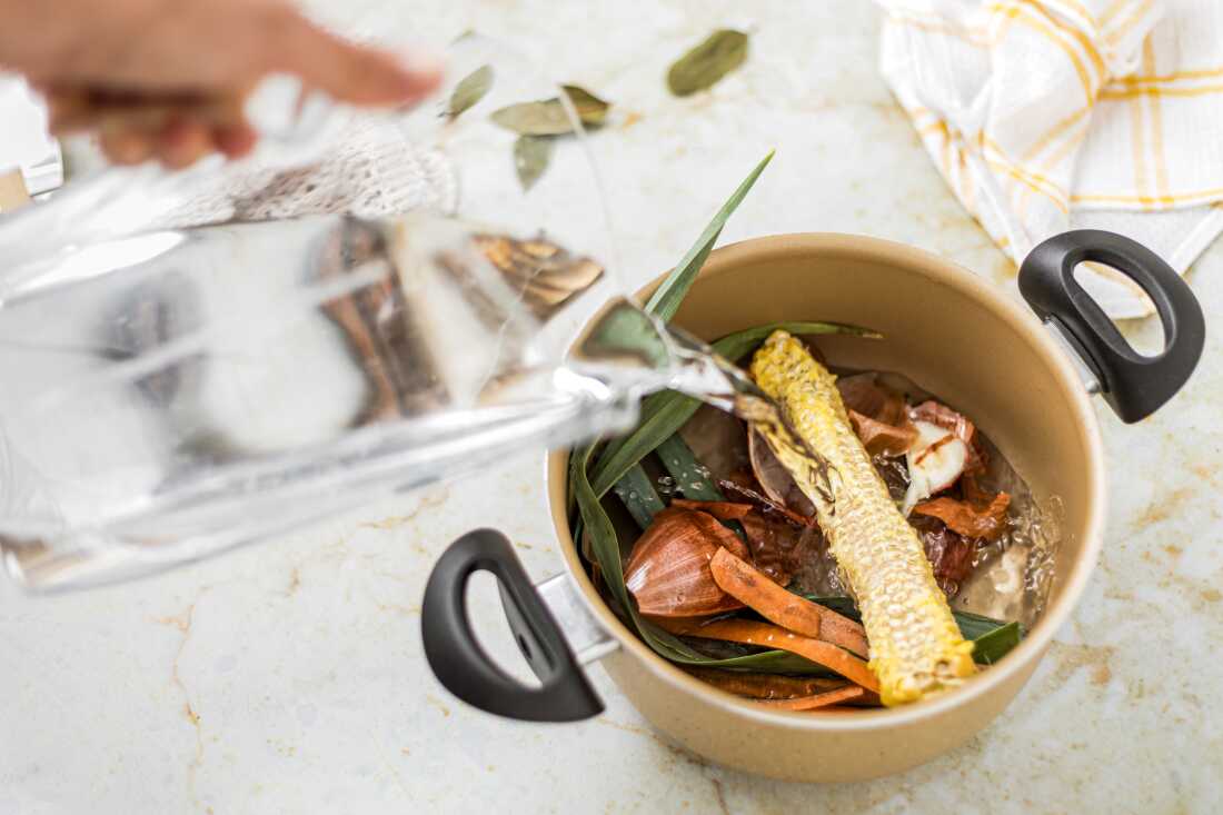 Cette photo montre un pot rempli de restes de légumes tels que des pelures de carottes et des pelures d'oignons. L'eau s'écoule dans le pot depuis un pichet transparent tenu par une main.