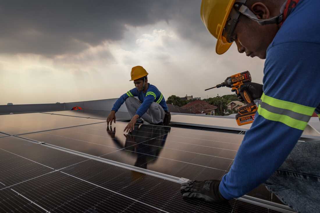 Des ouvriers portant des casques de sécurité installent des panneaux solaires sur le toit d’une maison sous un ciel rempli de nuages ​​gris.