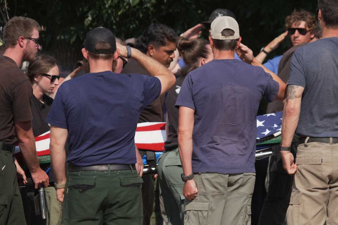 Plusieurs personnes saluent le corps d'un pompier tué tandis que d'autres portent le corps sur une civière. Un drapeau américain est drapé sur le corps.