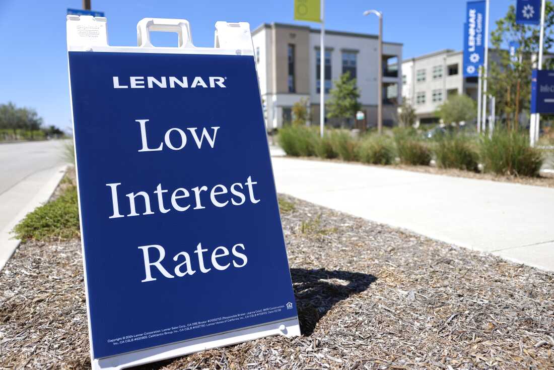 Un panneau en A indique "Lennar" et "Des taux d’intérêt bas" dessus. Les lettres sont blanches et sur fond bleu marine. Le panneau est placé sur une bande de terrain entre un trottoir et une bordure de rue.