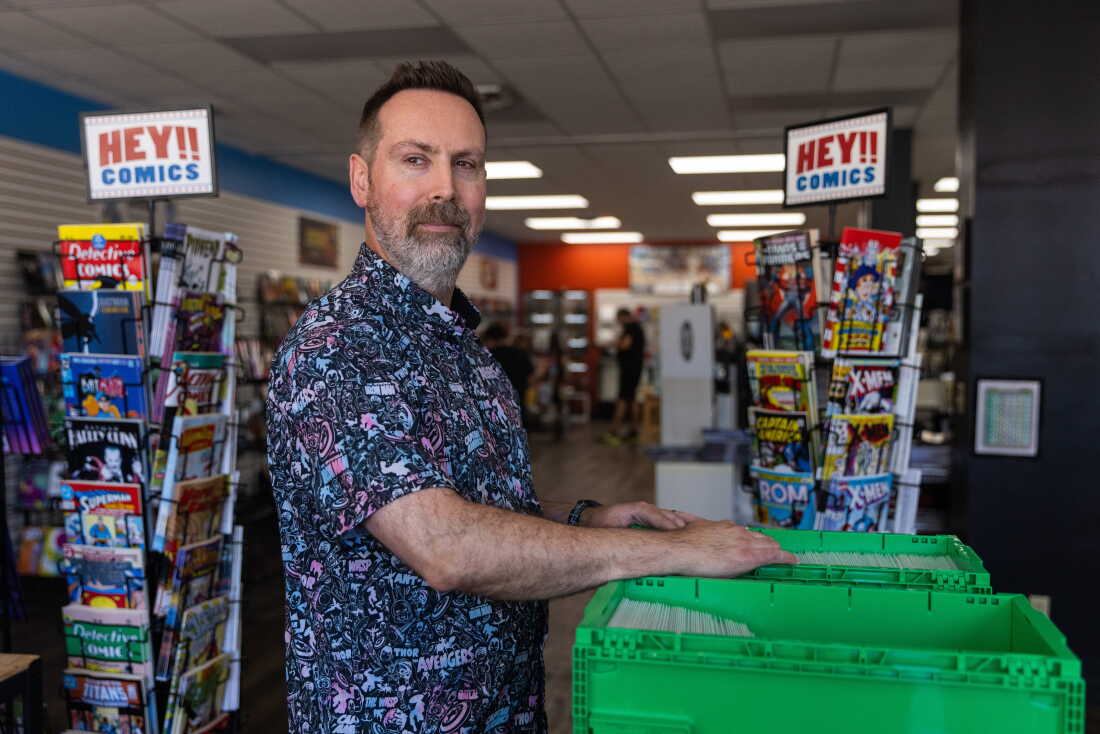 Marc Bowker, propriétaire d'Alter Ego Comics à Lima, Ohio, feuillette un bac vert rempli de bandes dessinées et de chaque côté de lui se trouvent deux étagères avec des pancartes indiquant : "Hé!! Bandes dessinées." Il a les cheveux bruns courts et une barbe grisonnante. Il porte une chemise boutonnée à motifs qui a "Vengeurs" écrit dessus avec des personnages Marvel tels que Iron Man et Captain America.