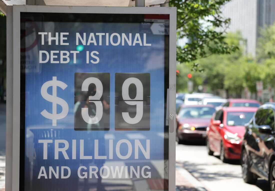 Un panneau sur le mur d’un arrêt de bus dit : "LA DETTE NATIONALE EST DE 39 000 MILLIARDS DE DOLLARS ET EN CROISSANCE." Les voitures passent sur une route du côté droit du cadre.