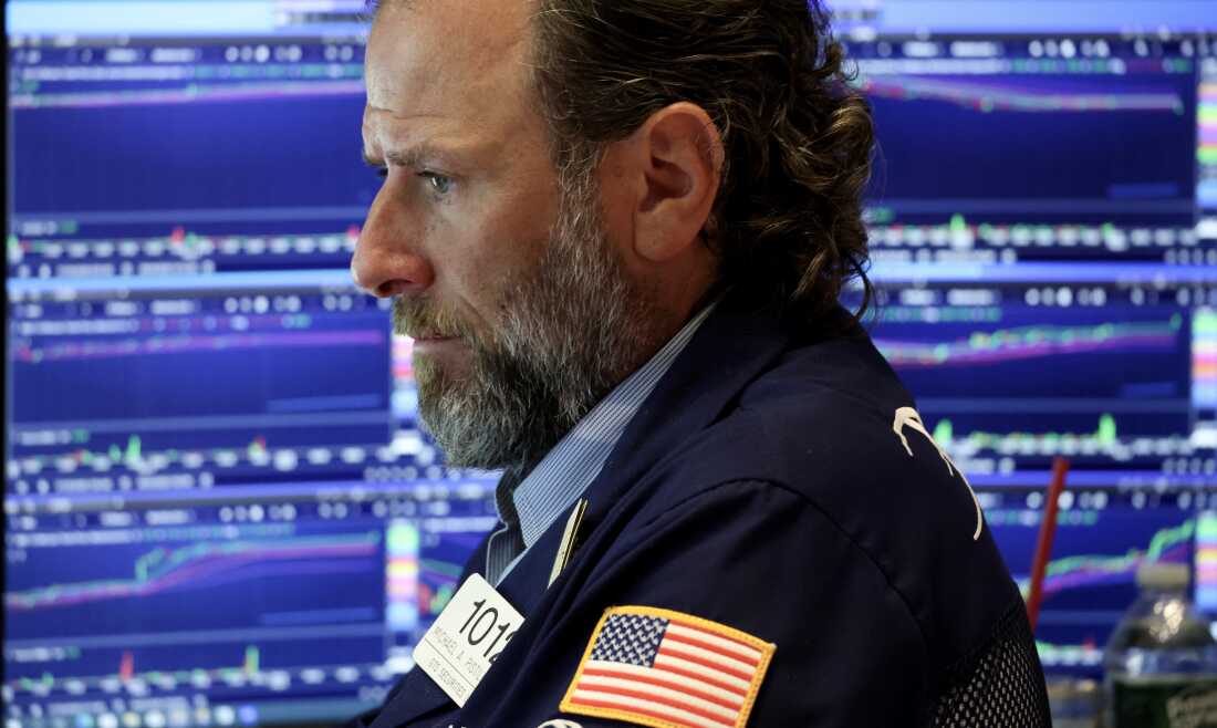 Un trader travaille sur le parquet d'une bourse. Il porte un blazer bleu marine et des écrans affichant des informations boursières sont derrière lui.