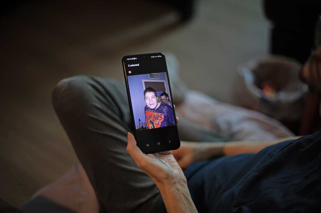 Debbie Willson tient son smartphone, qui affiche une photo de son fils, Adam Willson, décédé à l'âge de 27 ans en 2024. Adam porte un T-shirt sur lequel est écrit : "Jason vit," avec une image du personnage du film d'horreur Jason tenant une hache au-dessus de sa tête.