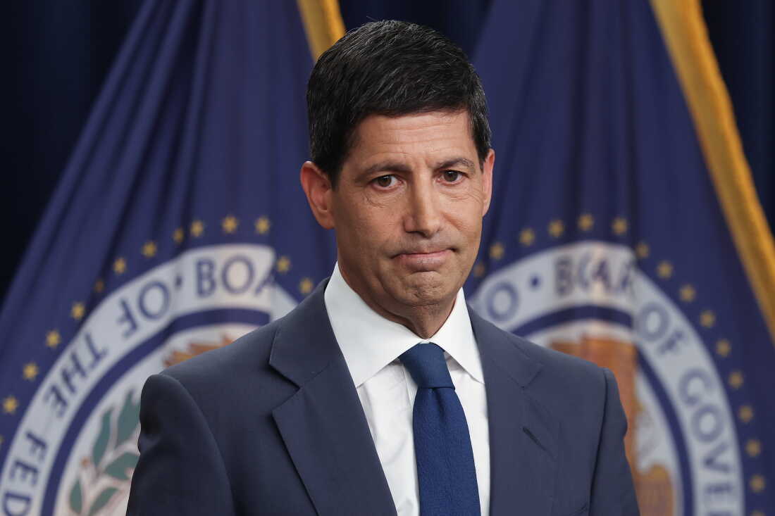 Cette photo montre le président de la Réserve fédérale, Kevin Warsh. Il porte un costume bleu, une chemise blanche et une cravate bleue. Derrière lui se trouvent deux drapeaux portant le sceau de la Réserve fédérale.