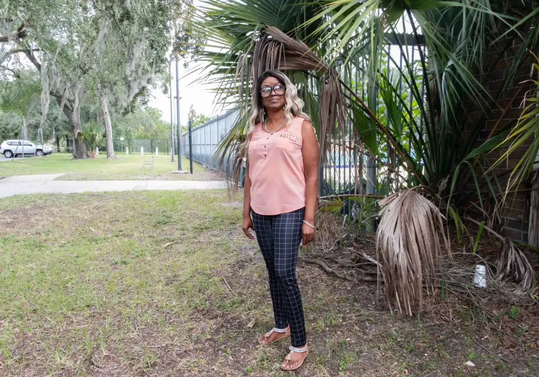 Rachel Dues, bénévole de Florida Rising, porte de grandes lunettes, un chemisier rose sans manches et un pantalon à carreaux. Elle a les cheveux noirs et blonds. Elle se tient à côté d’une clôture et d’une grande plante. 