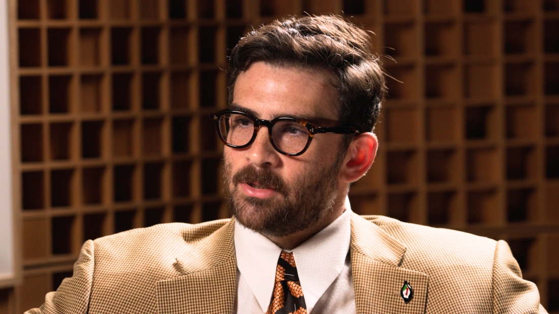 Hasan Piker est vu des épaules vers le haut, portant une veste de costume marron clair. 