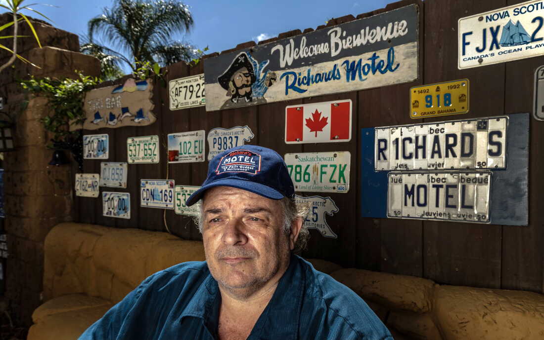 Richard Clavet est assis à l'extérieur à Hollywood, en Floride, le 27 mars 2025. Il porte une chemise bleue et une casquette de baseball bleue sur laquelle est écrit : "Motel de Richard" dessus. Derrière lui se trouve un panneau d'affichage en bois sur lequel sont accrochées plusieurs plaques d'immatriculation internationales. Les plaques d'immatriculation proviennent d'endroits tels que la Saskatchewan, la Nouvelle-Écosse et Grand Bahama.